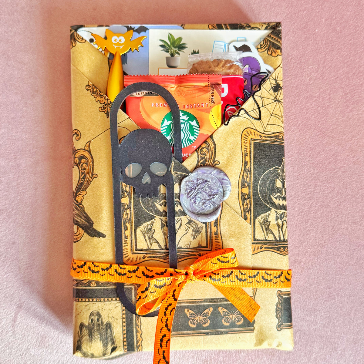 A Halloween-themed gift box wrapped in vintage-style brown paper with spooky illustrations, decorated with an orange ribbon, a black skull bookmark, a silver wax seal, and various small treats including a Starbucks coffee bag and a purple candy.