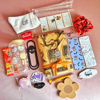 A flat-lay arrangement of Halloween-themed stationery and accessories on a dusty pink background, including a patterned notepad, a skull bookmark, a Biscuit Biscuit chocolate bar, a clear jar with heart stickers, a black hair clip, a red scrunchie, and other cute spooky items.
